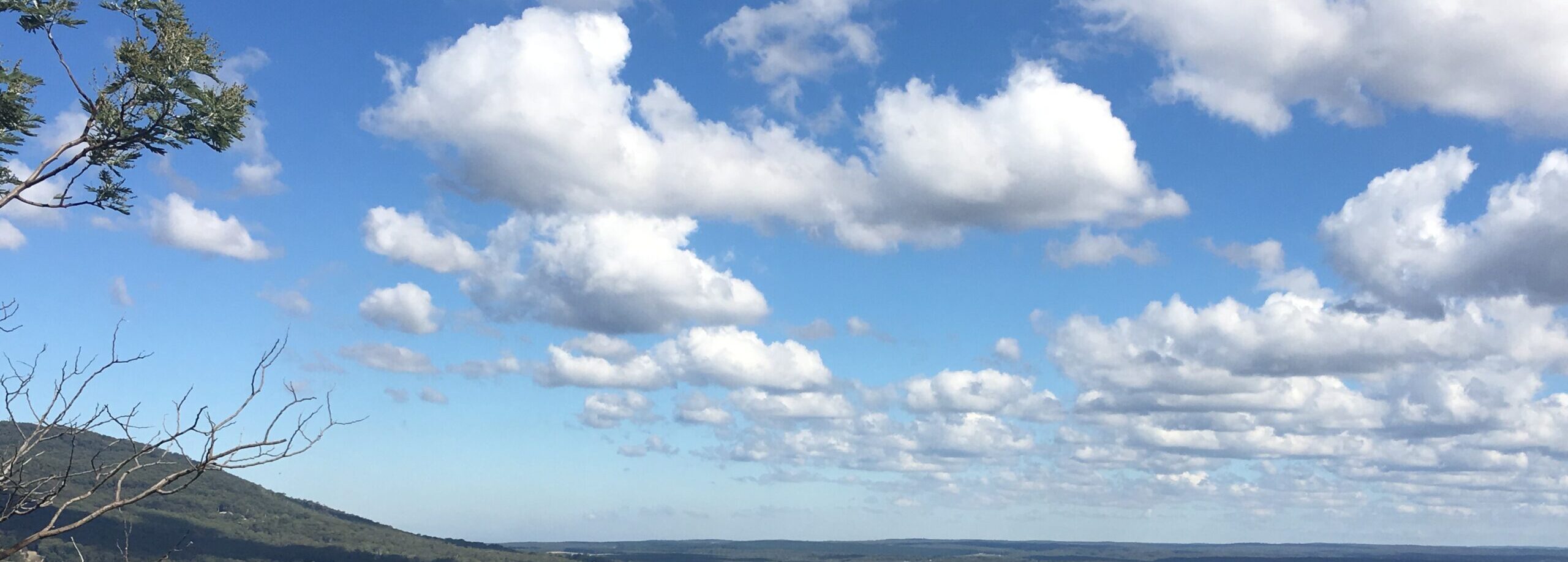 A blue sky filled with white, fluffy clouds over a landscape with trees and distant hills.